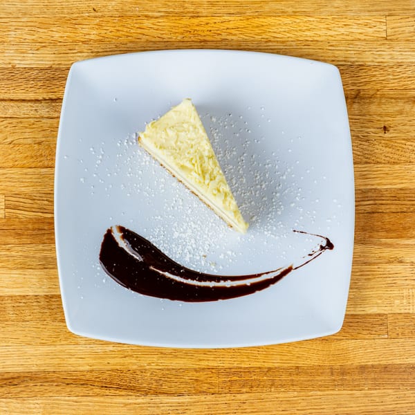 A slice of white chocolate cheesecake with powdered sugar and chocolate drizzle on a white plate.